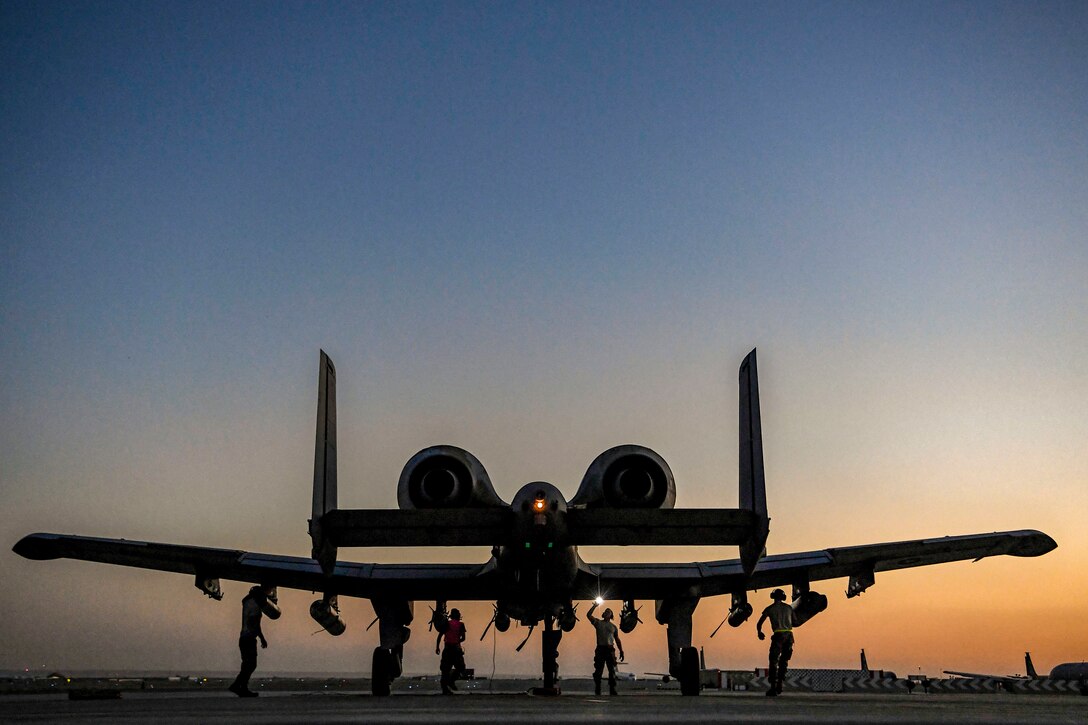 Four airmen, shown in silhouette, stand and work beneath the wings of an aircraft on a flightline.
