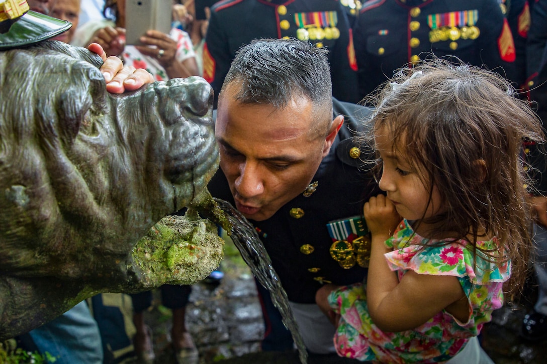 A Marine holds a girl while drinking water from a fountain shaped like a dog's head.