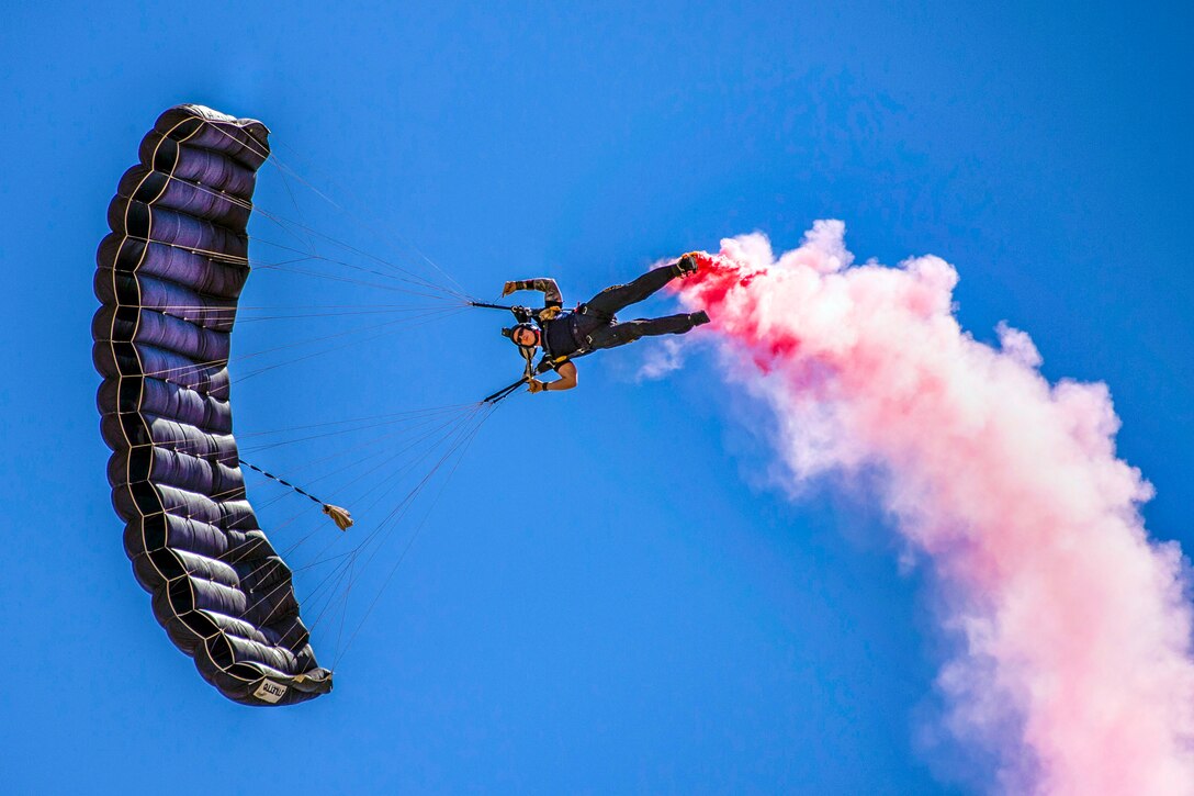 A parachutist floats sideways in blue sky while a pink cloud trails from his feet.
