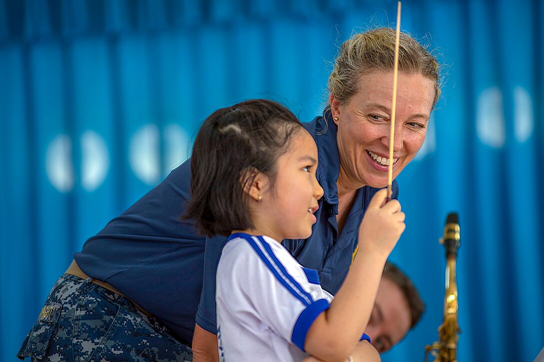 A little girl holds up a conductor's baton as a sailor looks on, framed by a bright blue background.
