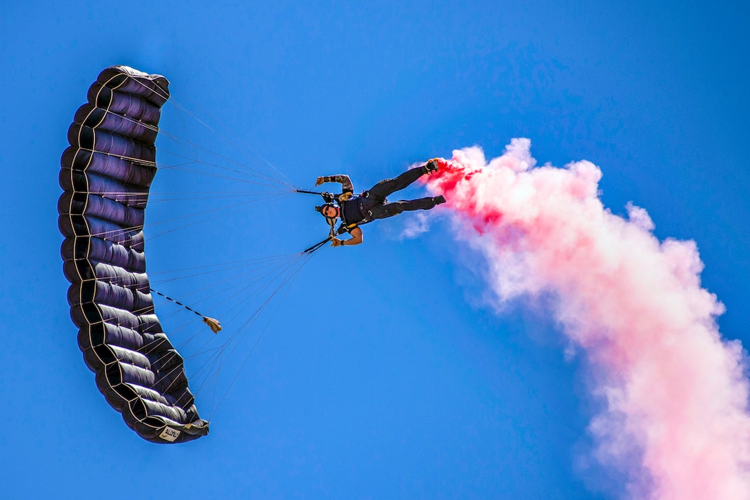 A parachutist floats sideways in blue sky while a pink cloud trails from his feet.