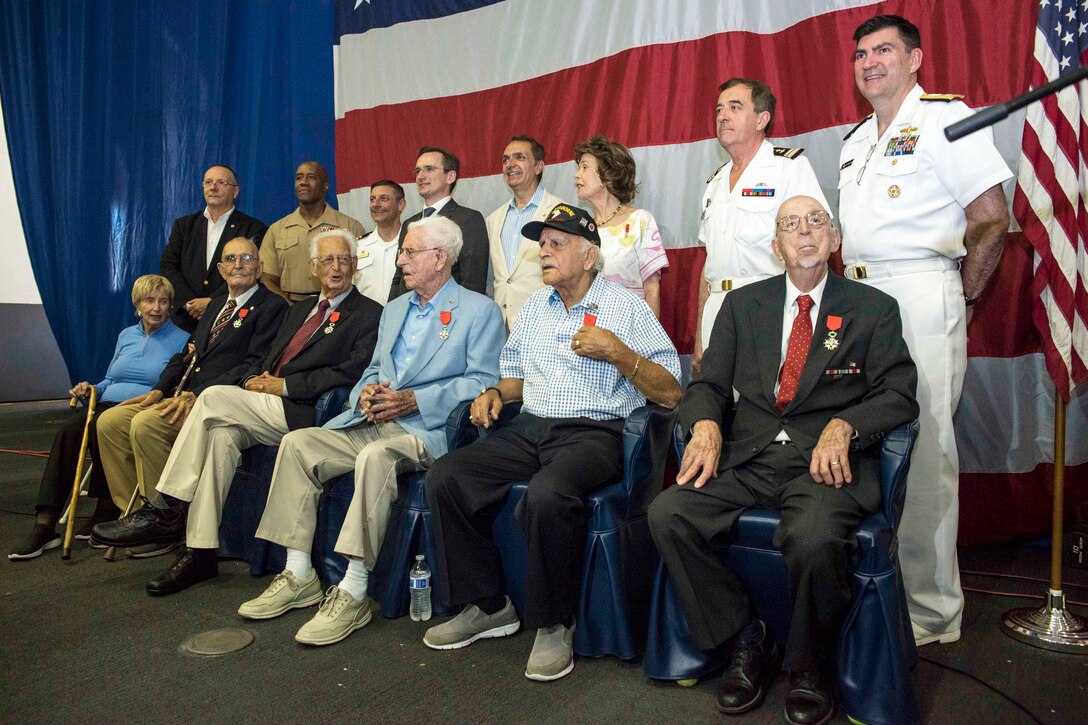 Fourteen people pose for a photo in two rows - one seated, one standing - in front of a U.S. flag.