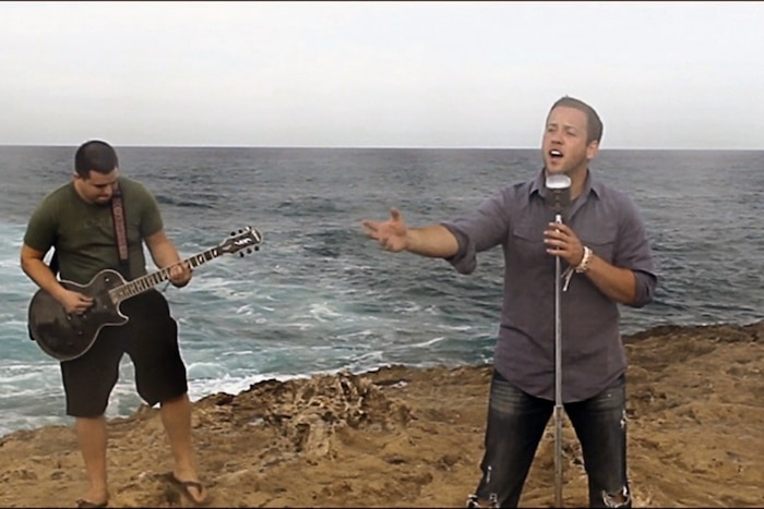A man sings into a microphone on a beach with another man playing the guitar next to him.