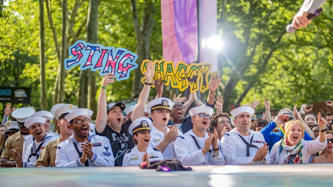 A performer's hand holds a microphone from a stage to a crowd of sailors and Marines, who sing and cheer.