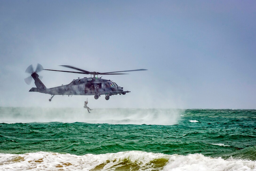 An airman hangs from a low-flying helicopter over green ocean water and sea spray.