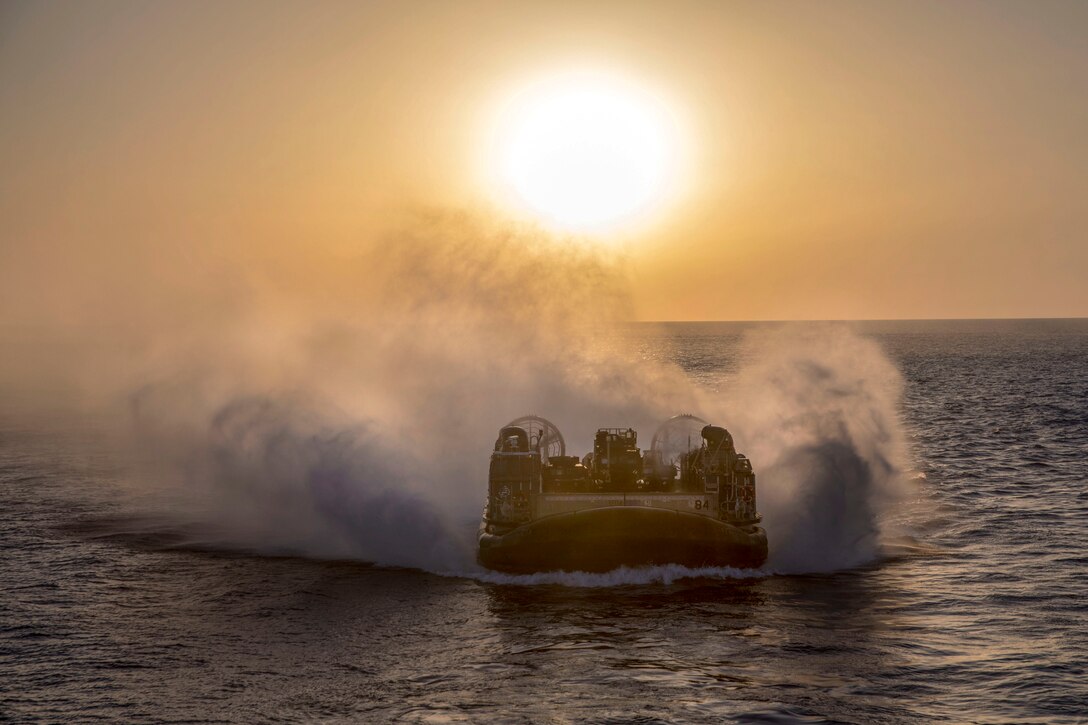 An air-cushioned landing craft sends up gusts of water while traveling in the sea, with a low sun in an orange sky behind it.
