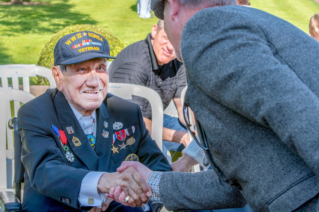 A World War II veteran wearing medals on his blazer shakes hands with someone with sitting at an outdoor ceremony.