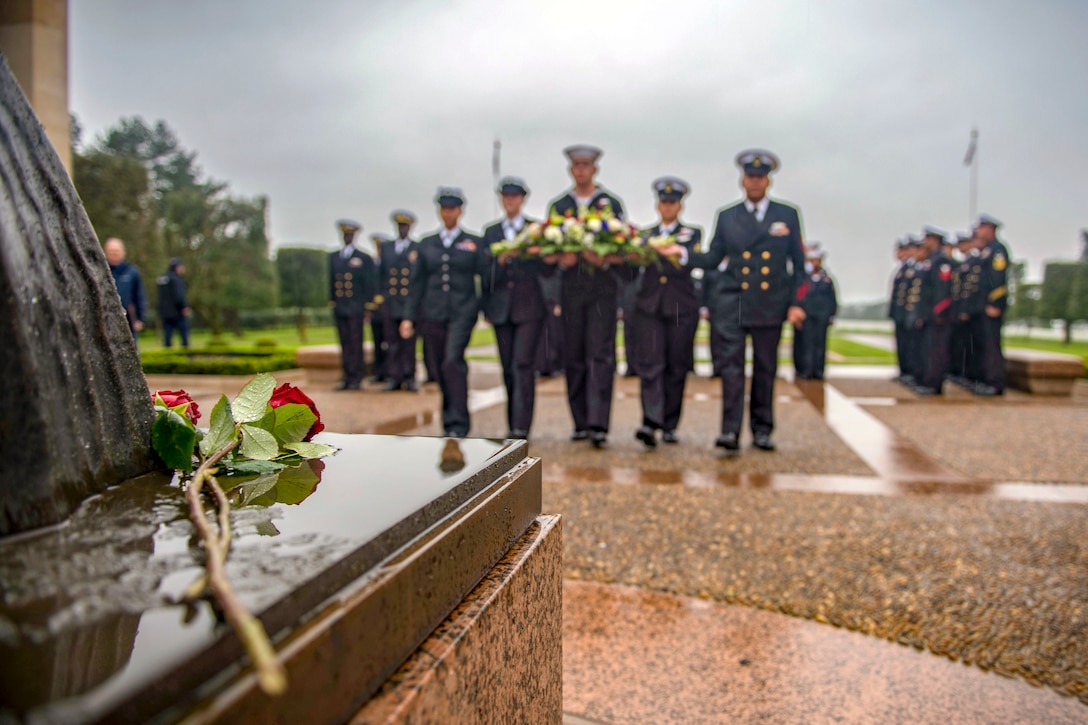 A rose sits at the base of a statue as sailors bearing a wreath walk toward it in formation.