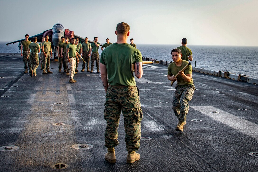 A Marine wielding a wooden dummy rifle creeps toward another Marine on a ship's flight deck, as a group observes.