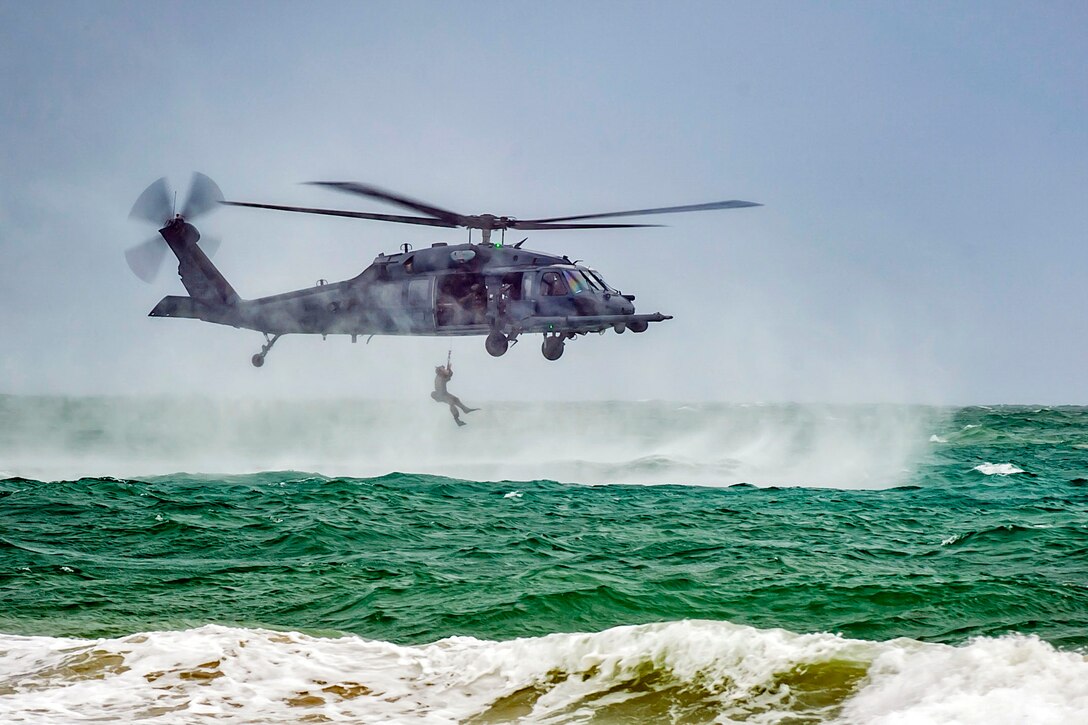 An airman hangs from a low-flying helicopter over green ocean water and sea spray.