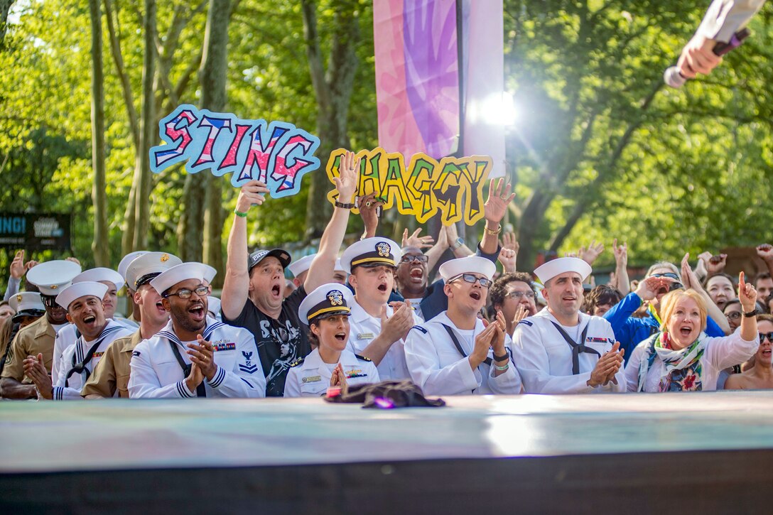 A performer's hand holds a microphone from a stage to a crowd of sailors and Marines, who sing and cheer.