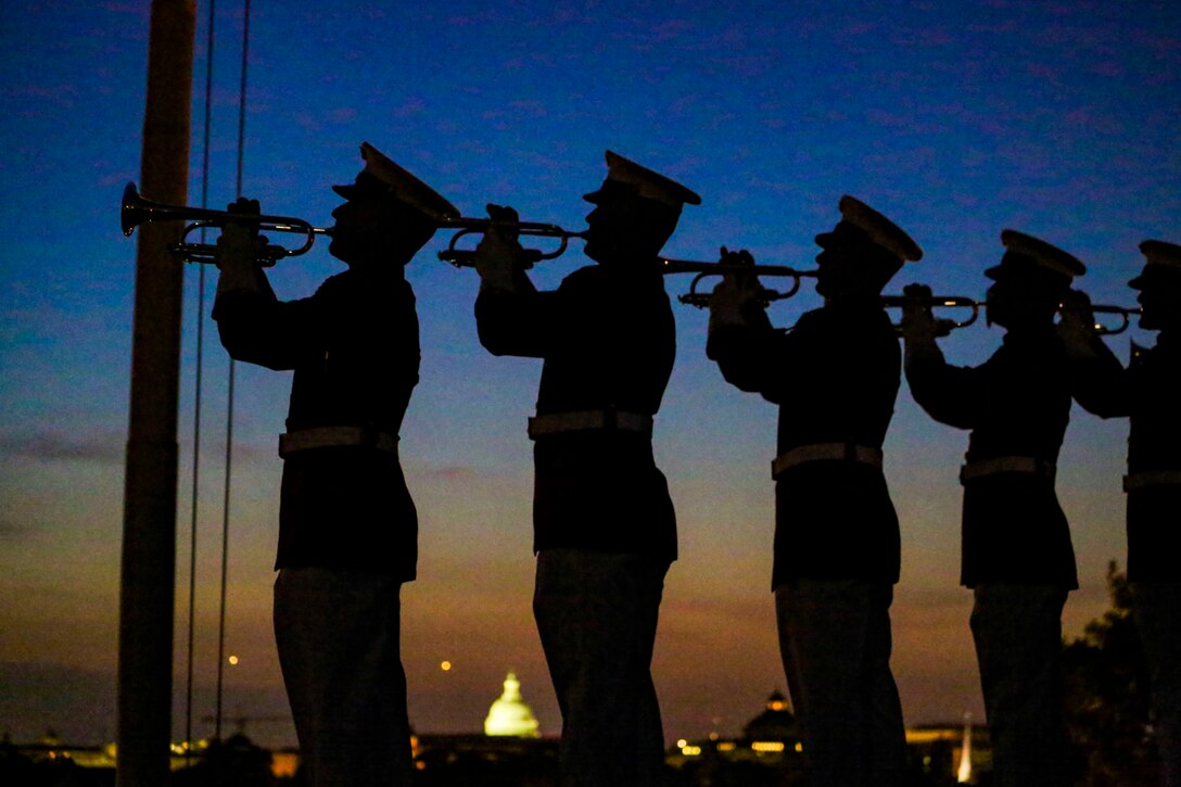 Marines, shown in silhouette, stand in a row and play bugles, against a deep blue and orange sky with the U.S. Capitol in the distance.