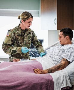 Caucasian female Officer Nurse 1LT Oskam standing bedside with male patient administering treatment inside a hospital.