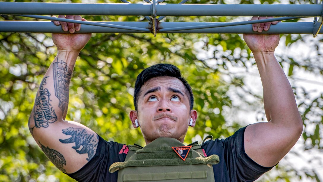 A sailor looks up while doing a pullup with trees in the background.