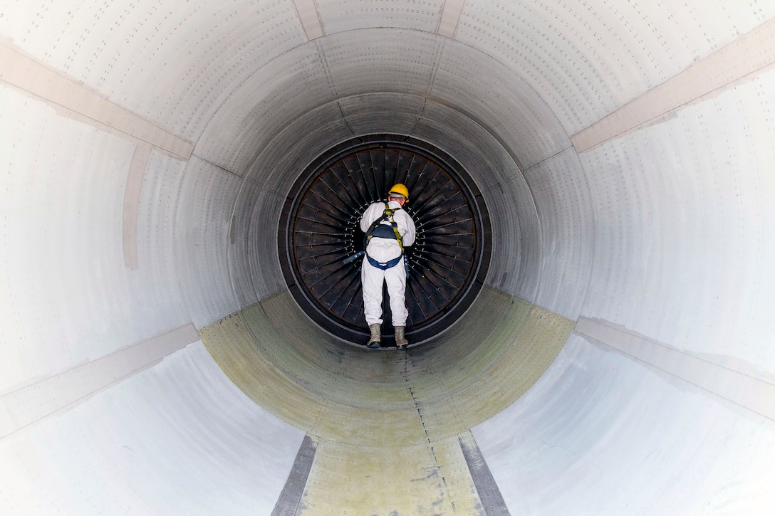 An airman in white works inside a white, tunnel-shaped structure on a circular object at its center.