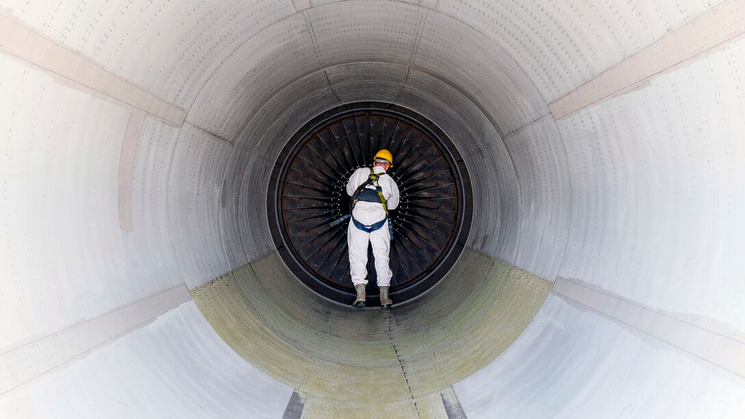 An airman in white works inside a white, tunnel-shaped structure on a circular object at its center.