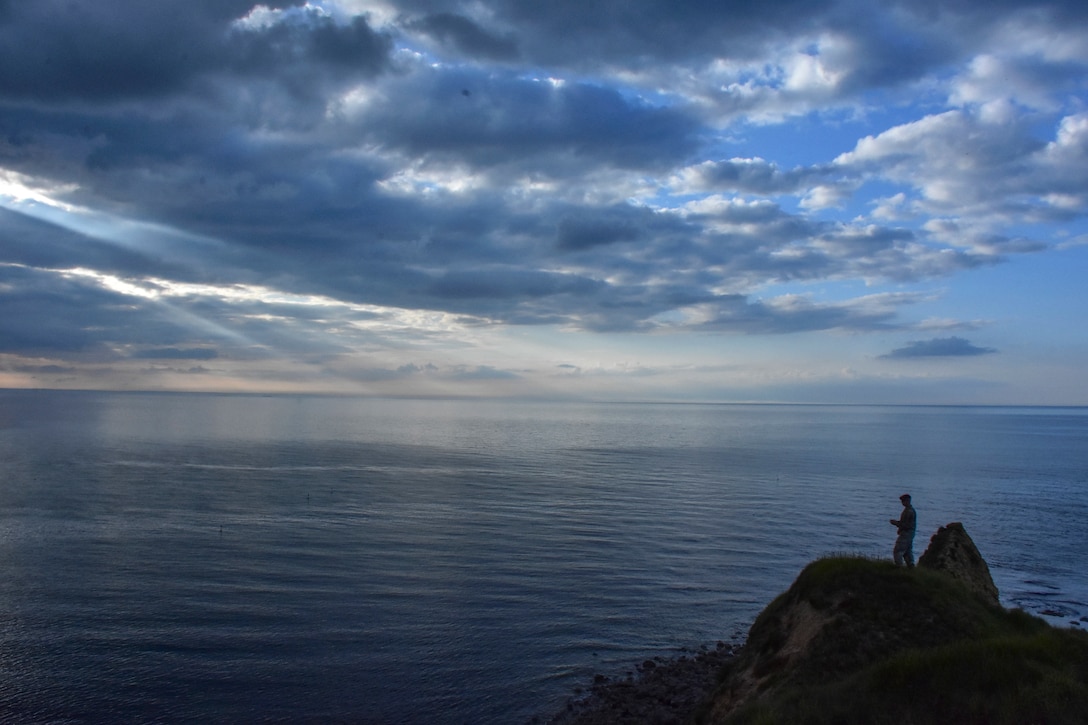 A soldier stands on a rocky promontory and looks out over a body of water below a low-lit, sun-streaked sky.