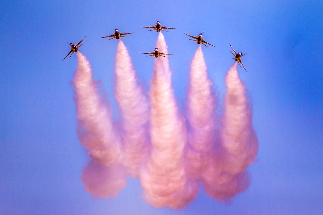 Pink plumes of smoke emanate from six jets flying in formation in a cloudless blue sky.
