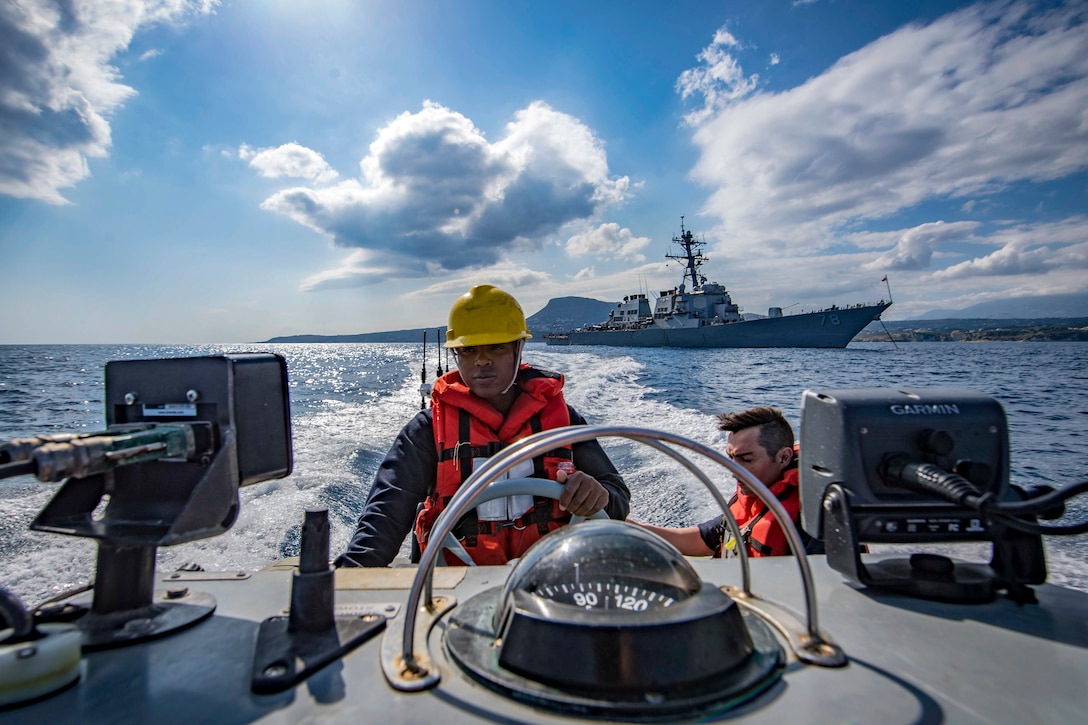 A sailor drives a boat, as a larger ship travels in the distance.