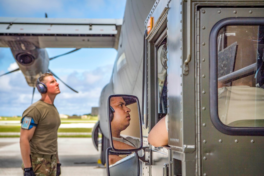 Two airmen stare in the same direction on a flightline, as one stands and one sits in a nearby truck, his face visible in the rear-view mirror.