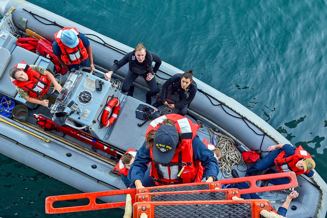 A sailor climbs down an orange ladder toward an inflatable boat, where seated sailors are looking up.