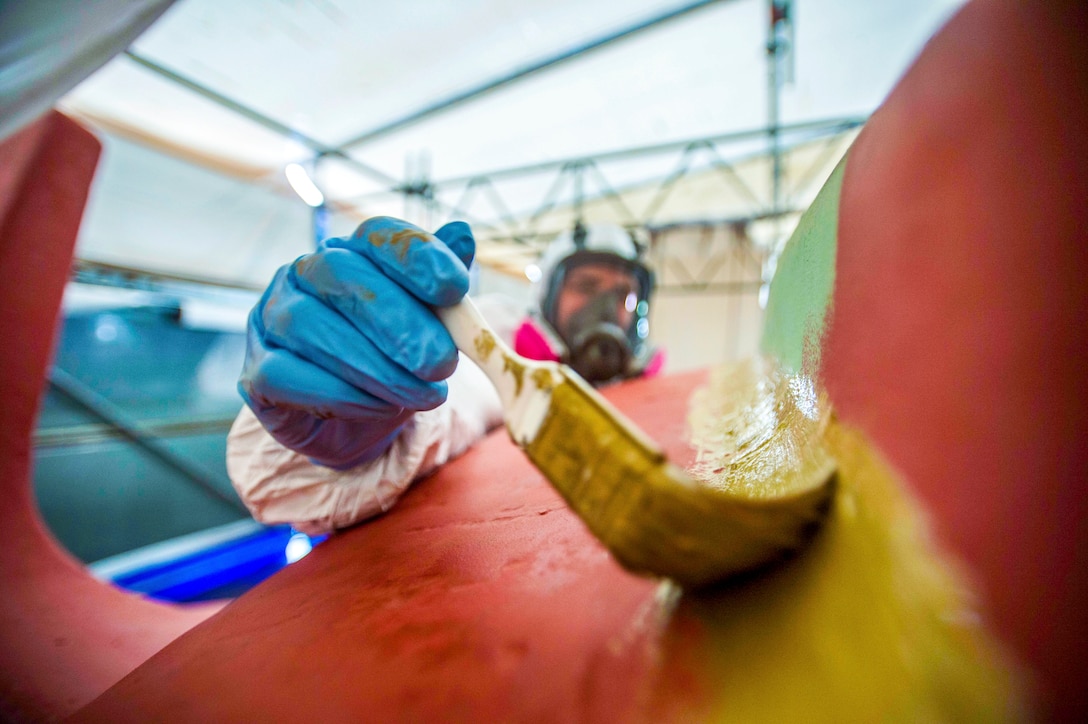 A sailor in white coveralls and blue gloves applies yellow paint to an anchor.