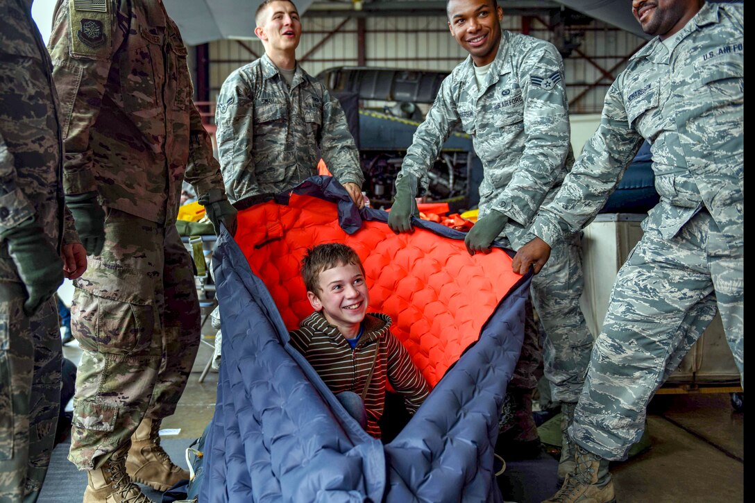 A smiling child sits in inside a quilted bag-type item being held up by smiling airmen.