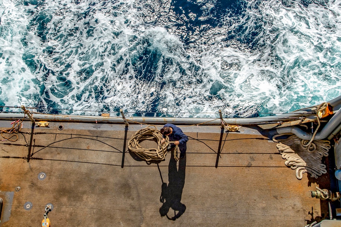 From above, a navy seaman coils a rope next to the railing of a ship.