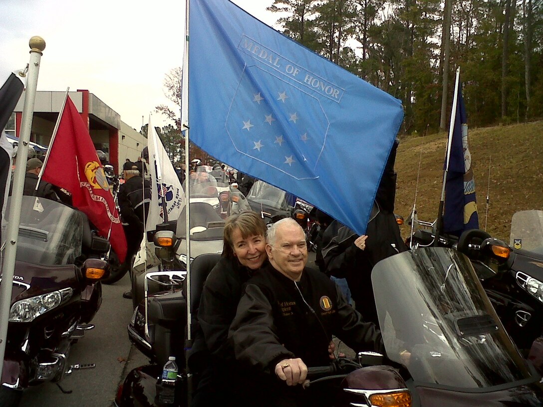 Baker and wife sit on motorcycle surrounded by other motorcycles.