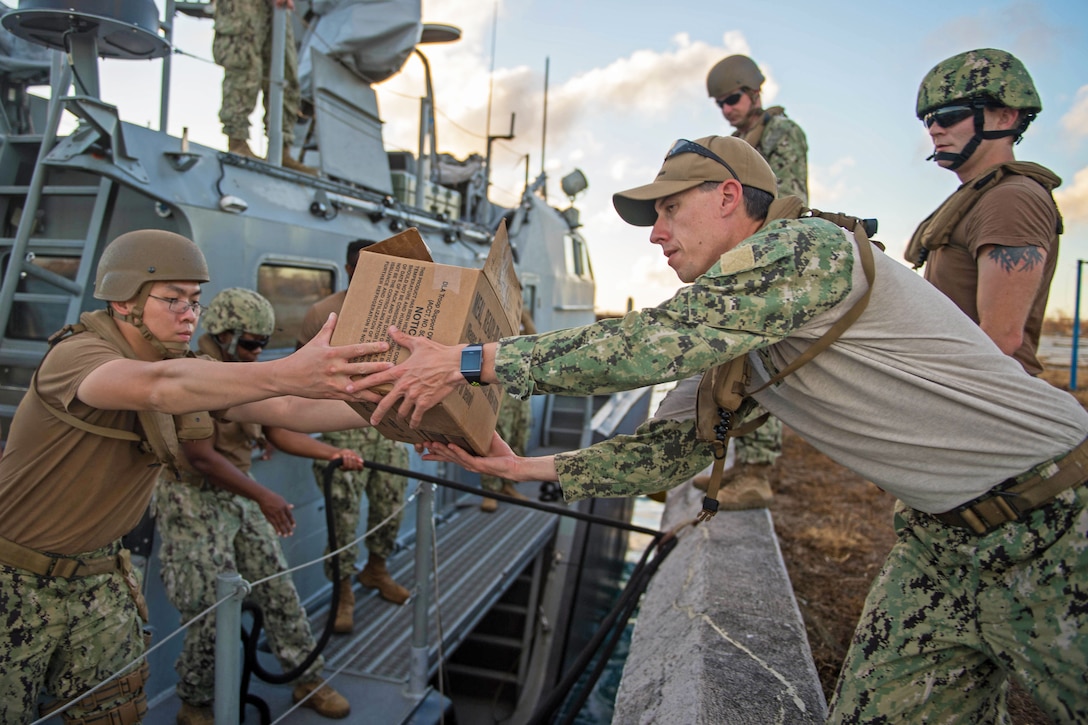 A sailor in a boat hands a box to another on the pier while others stand nearby.