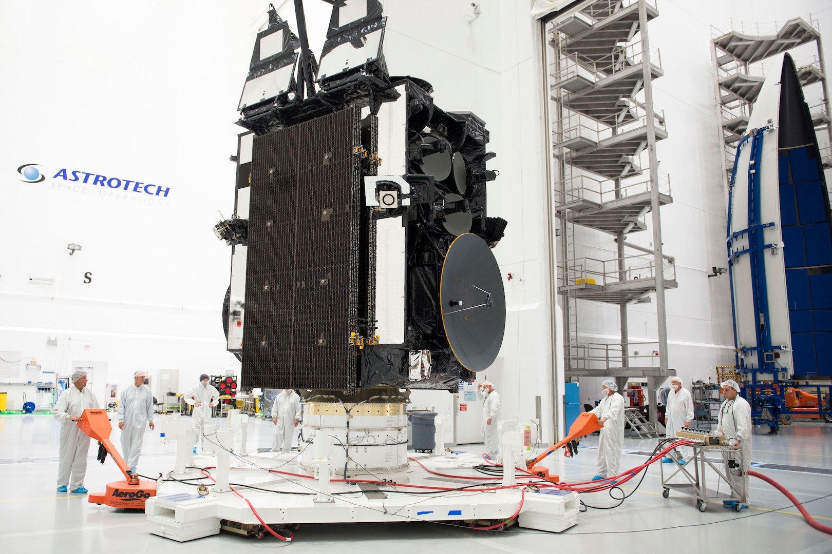 Large satellite about to be enclosed in two half-shell rocket panels, in a white room surrounded by people in white coveralls and head coverings.