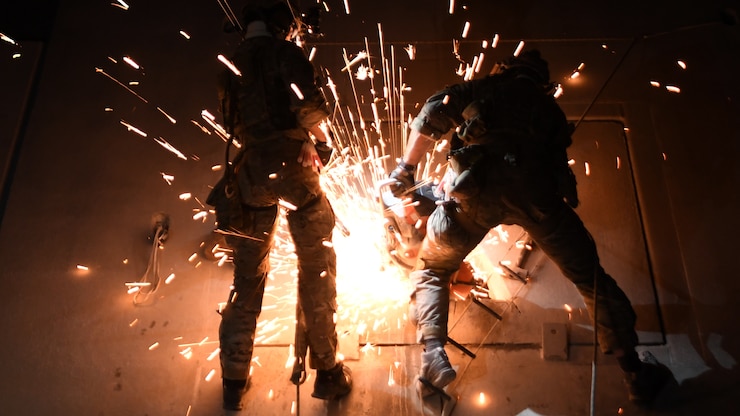 A pair of Special Tactics Operators hang suspended by harnesses in the dark and use a cutting wheel to cut through a metal hatch to make an opening.