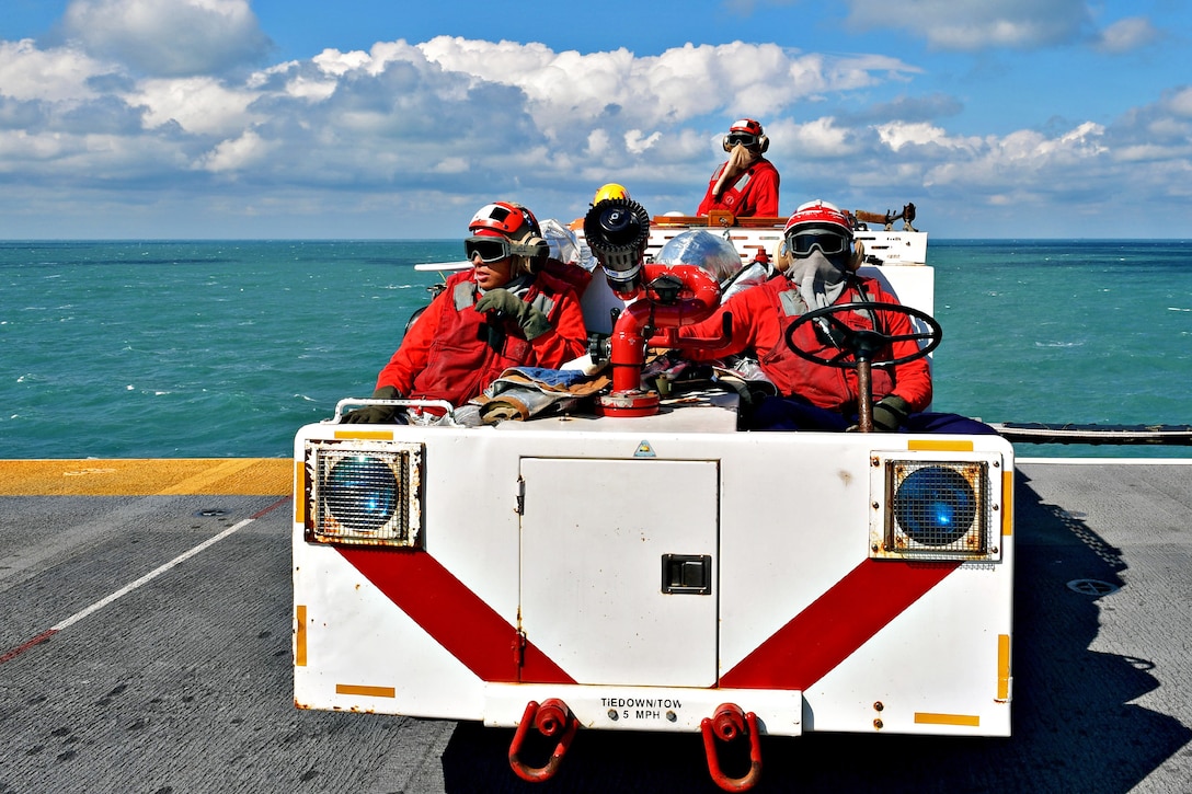 Three sailors keep watch from a cart on the flight deck of a ship in the sea.