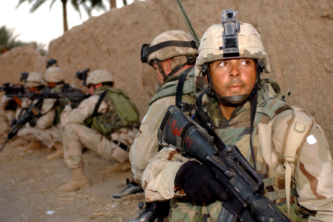 A soldier in a helmet and camo holds a rifle while standing by a mud wall in a line with other troops.