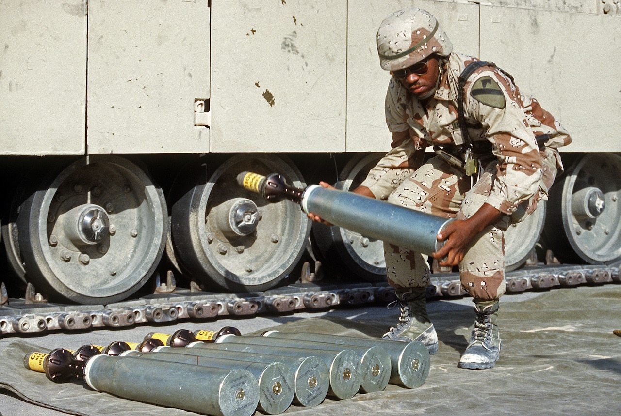 A service member lifts a metal round in front of a wheeled vehicle.