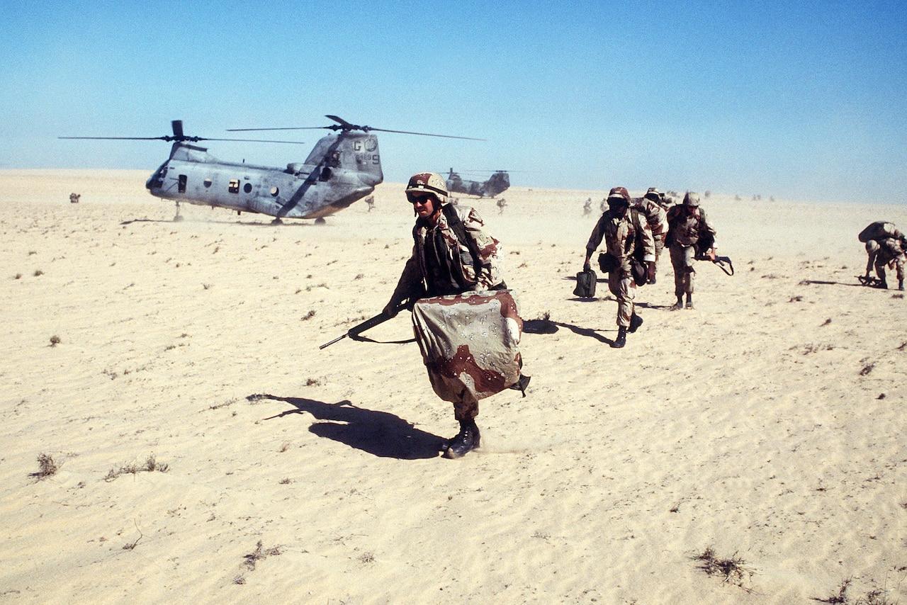 Marines run over sandy terrain near a helicopter.