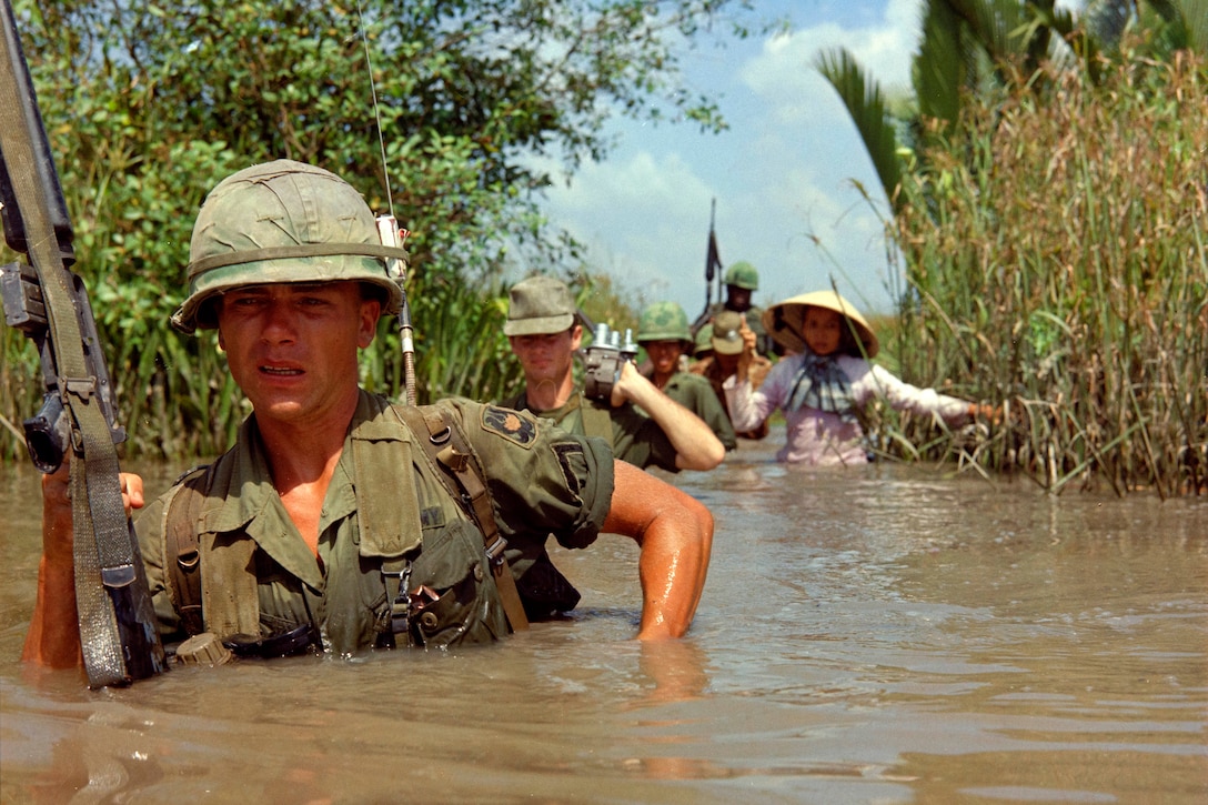 A soldier carrying a rifle leads others through chest-deep muddy water.