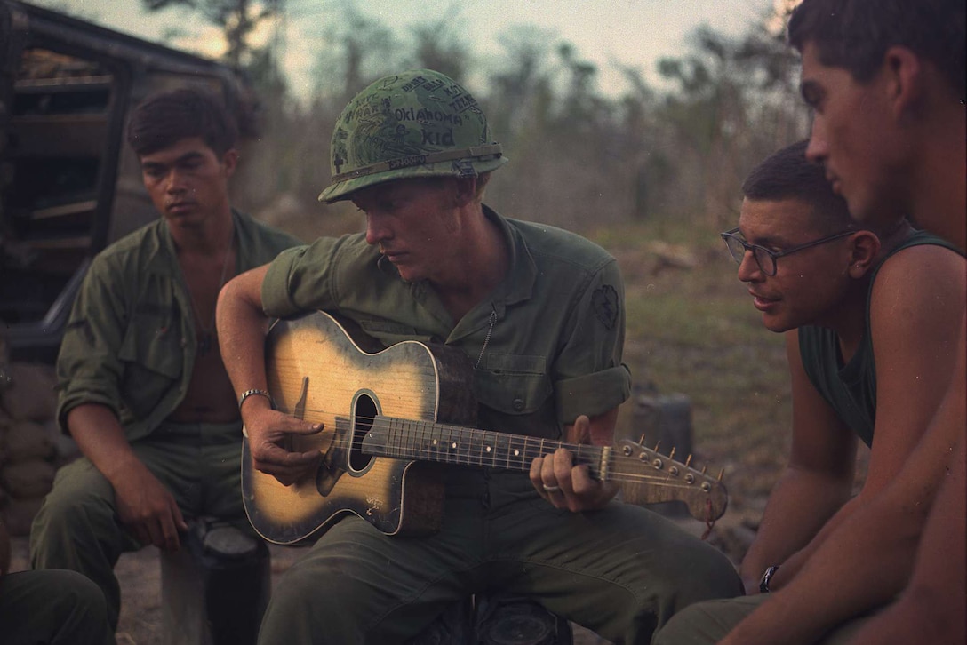 A service member sits and strums a guitar outside as others sit by him and watch.