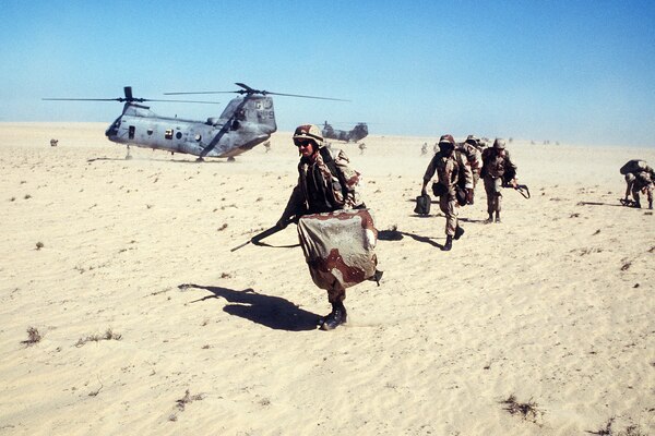 Marines run over sandy terrain near a helicopter.