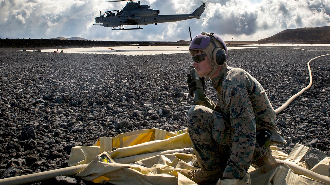 A Marine is on the radio while adjusting a fuel line running into the background for a landing helicopter.