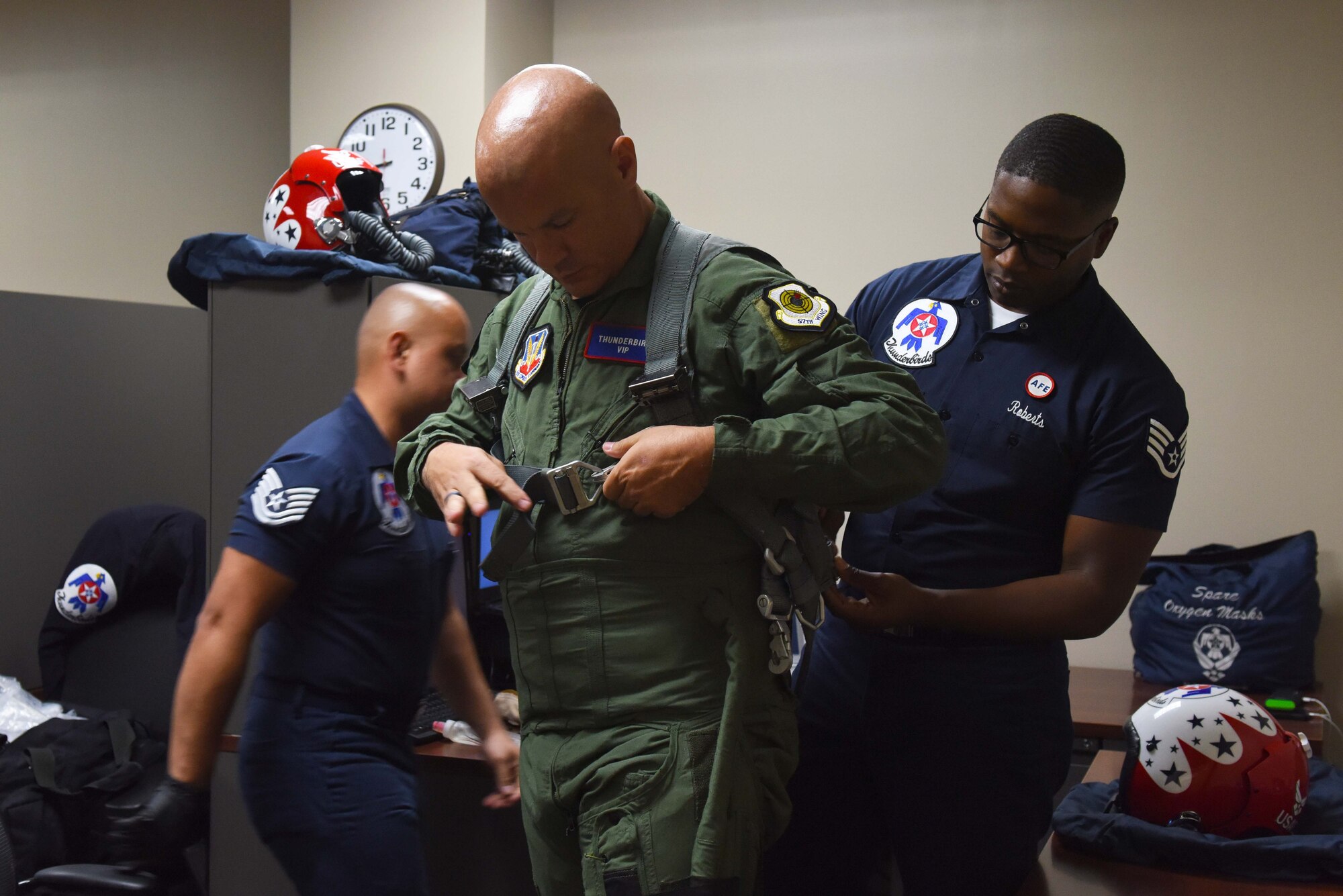 A man in a green flight suit is helped by another man on the right wearing a blue uniform. The man in the green suit is getting fitted with a sage green parachute.