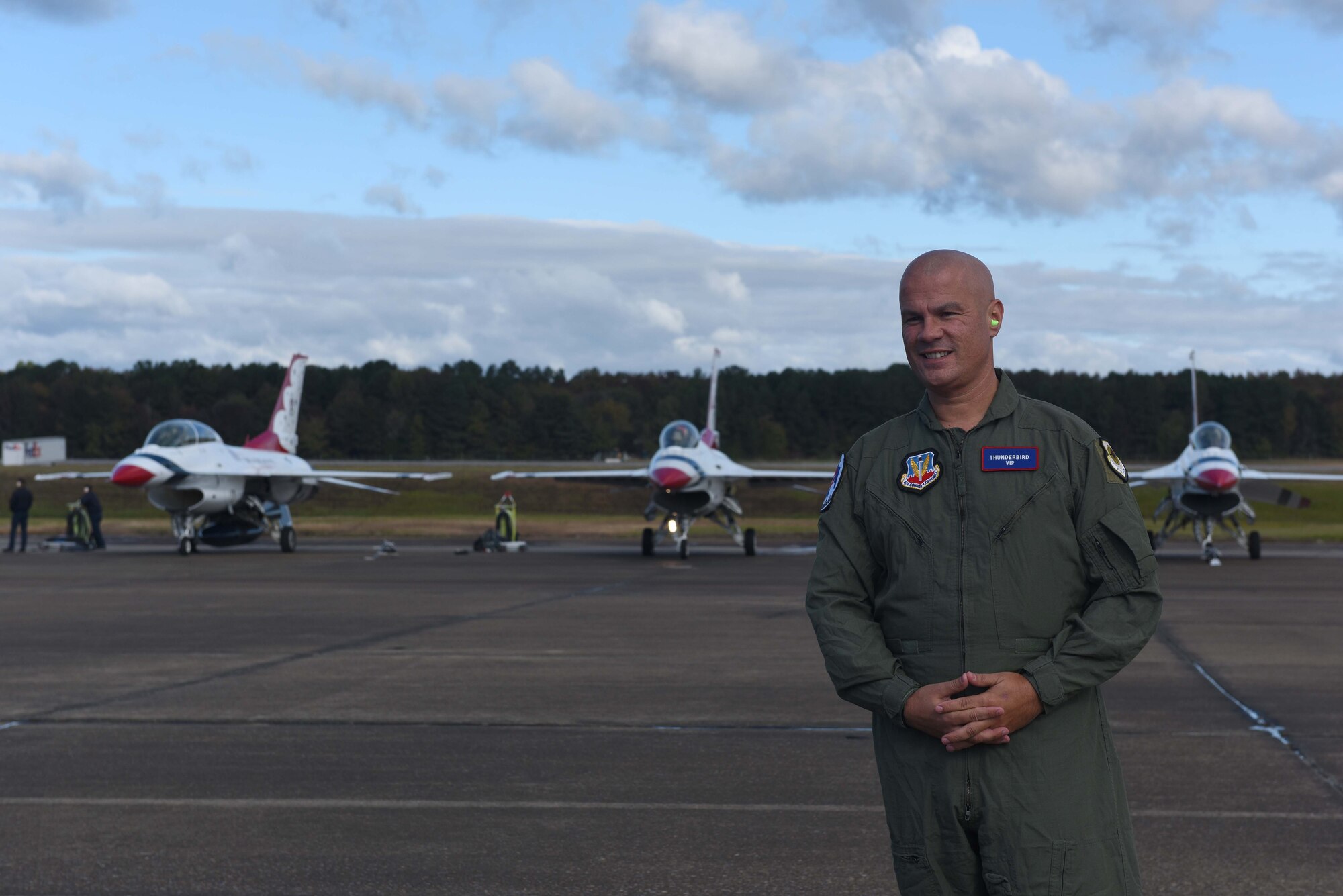 A man in a green flight suit stands before three white aircraft on a flightline.
