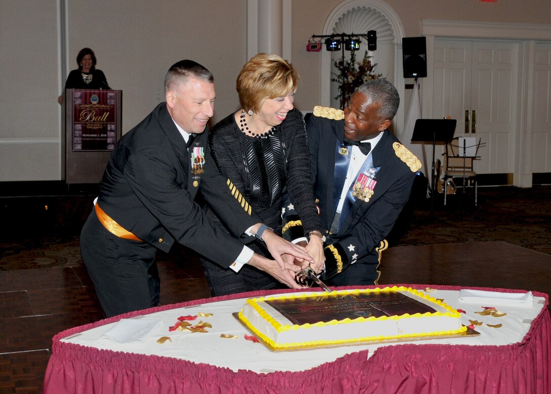 Left to right, Senior Enlisted Leader Navy Command Master Chief Shaun Brahmsteadt,  Under Secretary of Defense for Acquisition and Sustainment Ellen Lord and DLA Director Army Lt. Gen. Darrell Williams cut the cake with a ceremonial sword as DLA Chief of Staff Kristin French, officiates from lectern in background.