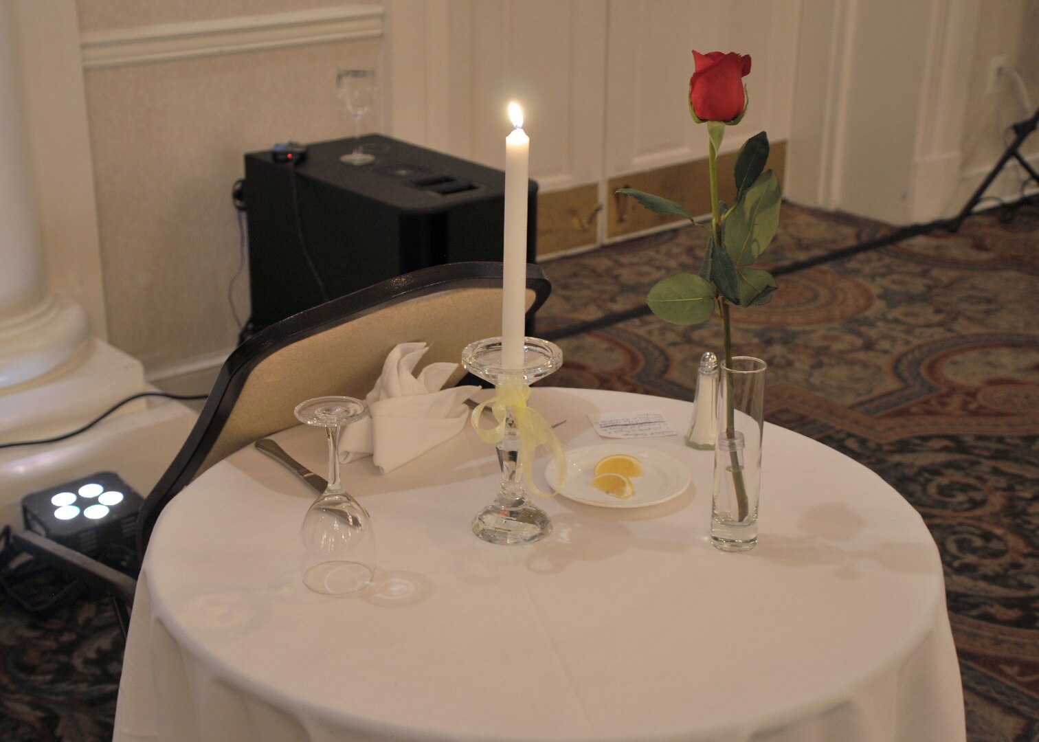 A chair is tipped up against a round table with white tablecloth, candle, single rose in a vase, an inverted glass and wedge of lemon on a plate. All symbolize the missing military and civilian members who cannot be present at the festivities.