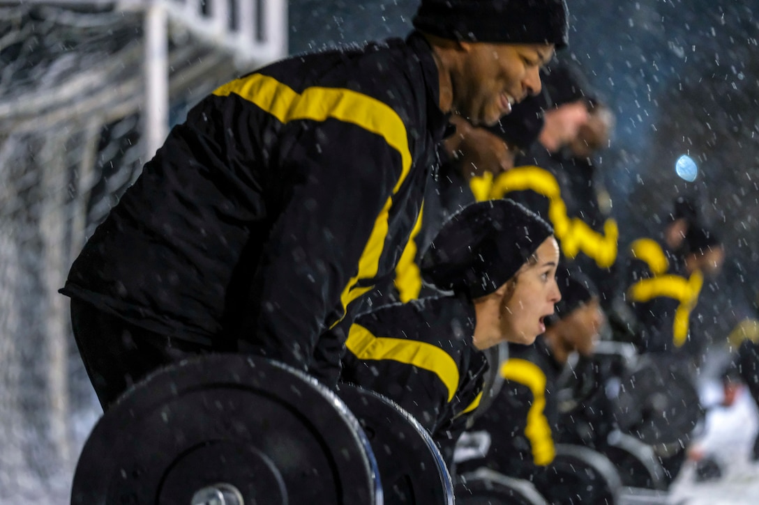 Soldiers in matching black and yellow warmup clothes lift weights in the snow.