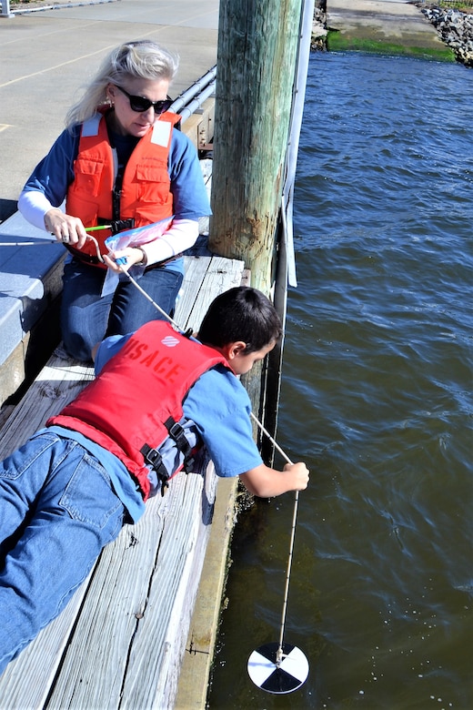 Fifth-grade boy for Norfolk Christian Schools is hanging a black and white disk from a rope off of a dock while his female teacher kneels beside him to help