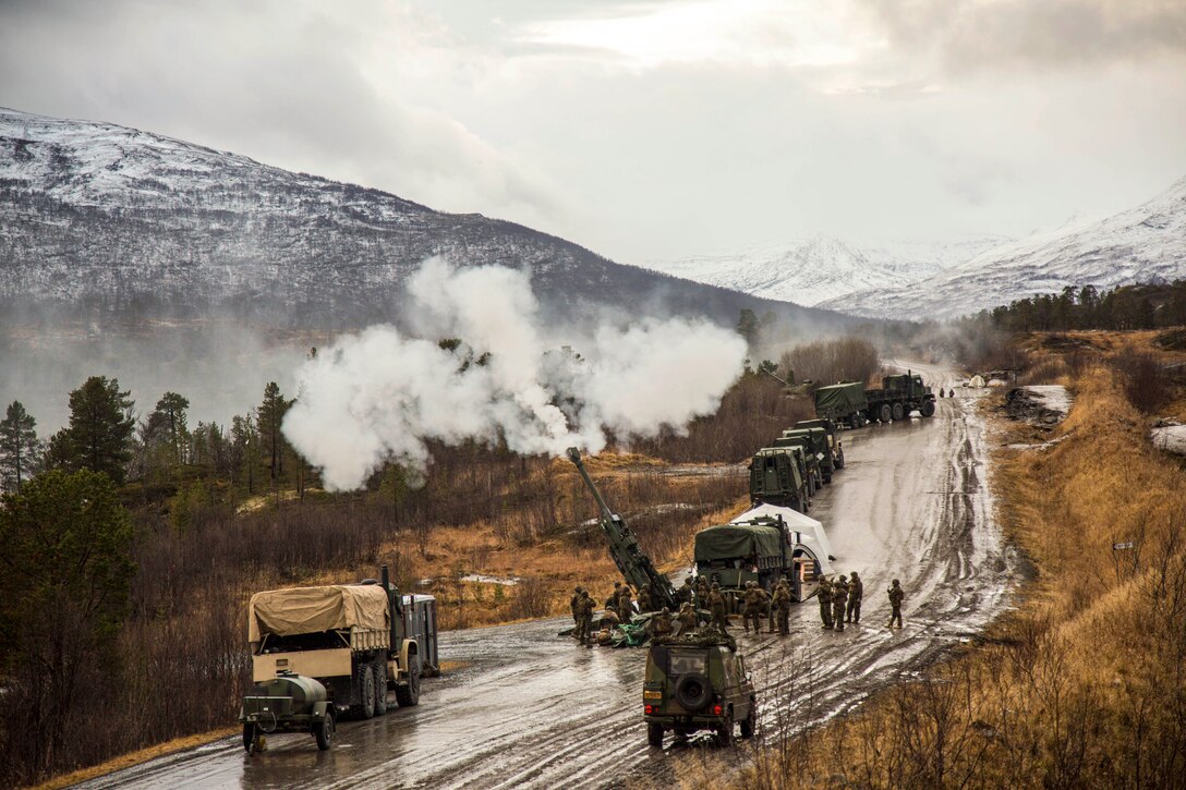 Troops fire a howitzer on a  road with snowy mountains in the distance.