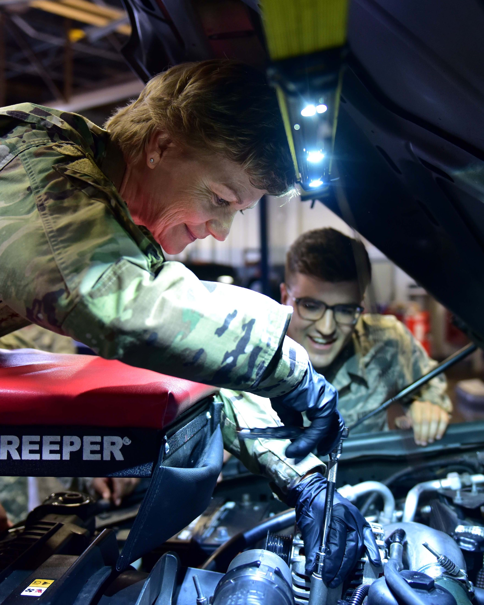 A woman wearing the operational camouflage pattern uniform changes a car alternator while an Airman in the back wearing the Airman Battle Uniform stands behind her while she changes it.