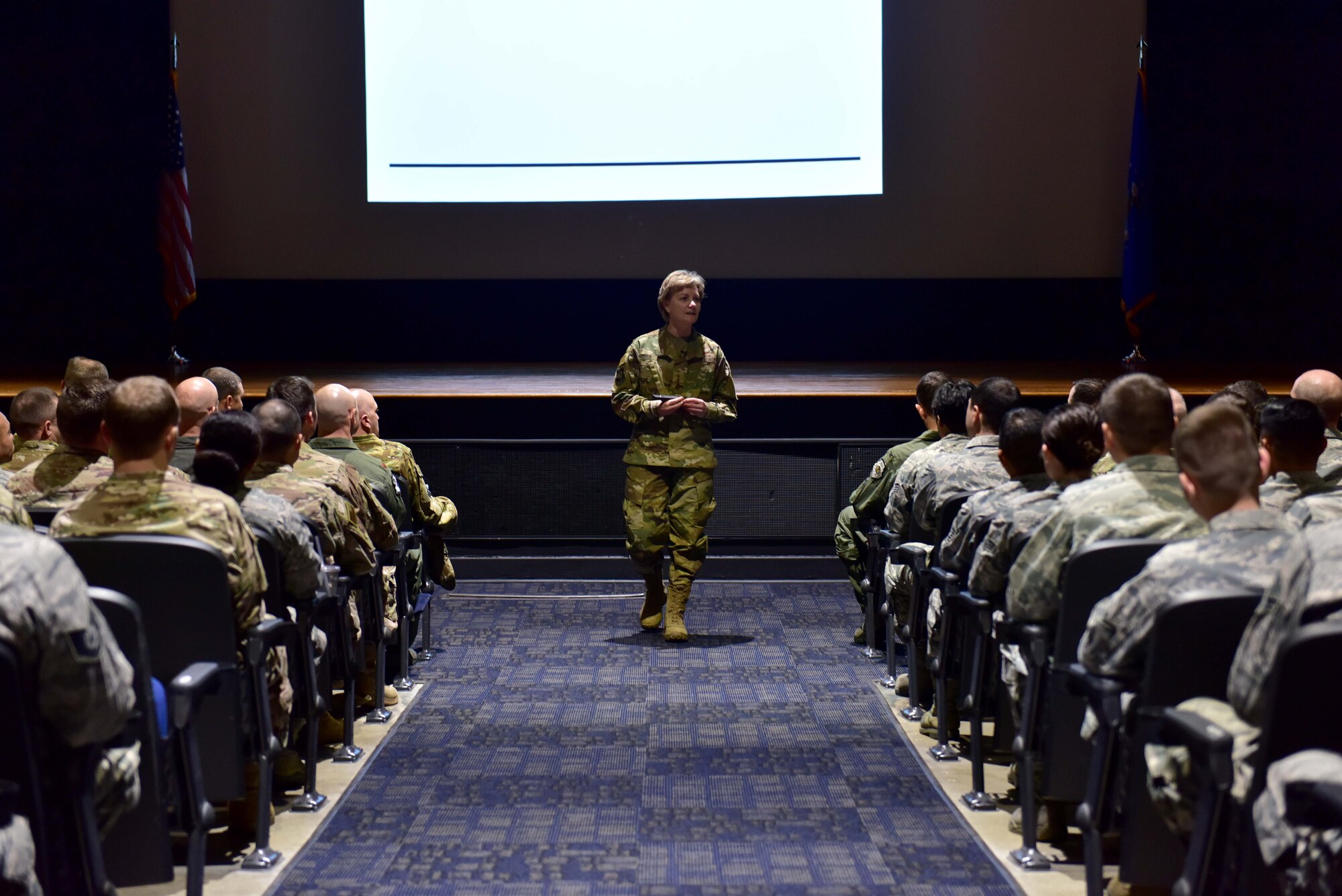 A woman wearing the operational camouflage pattern uniform speaks to a room full of Airmen.