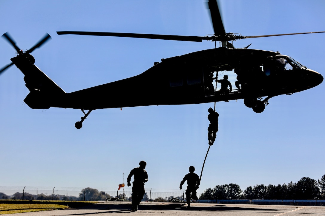 An Army Ranger slides down a rope out of a helicopter as two fellow Rangers run on a flightline.